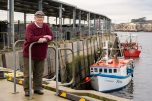 Terry McDermott standing on North Shields fish quay with fishing trawlers moored in the background.